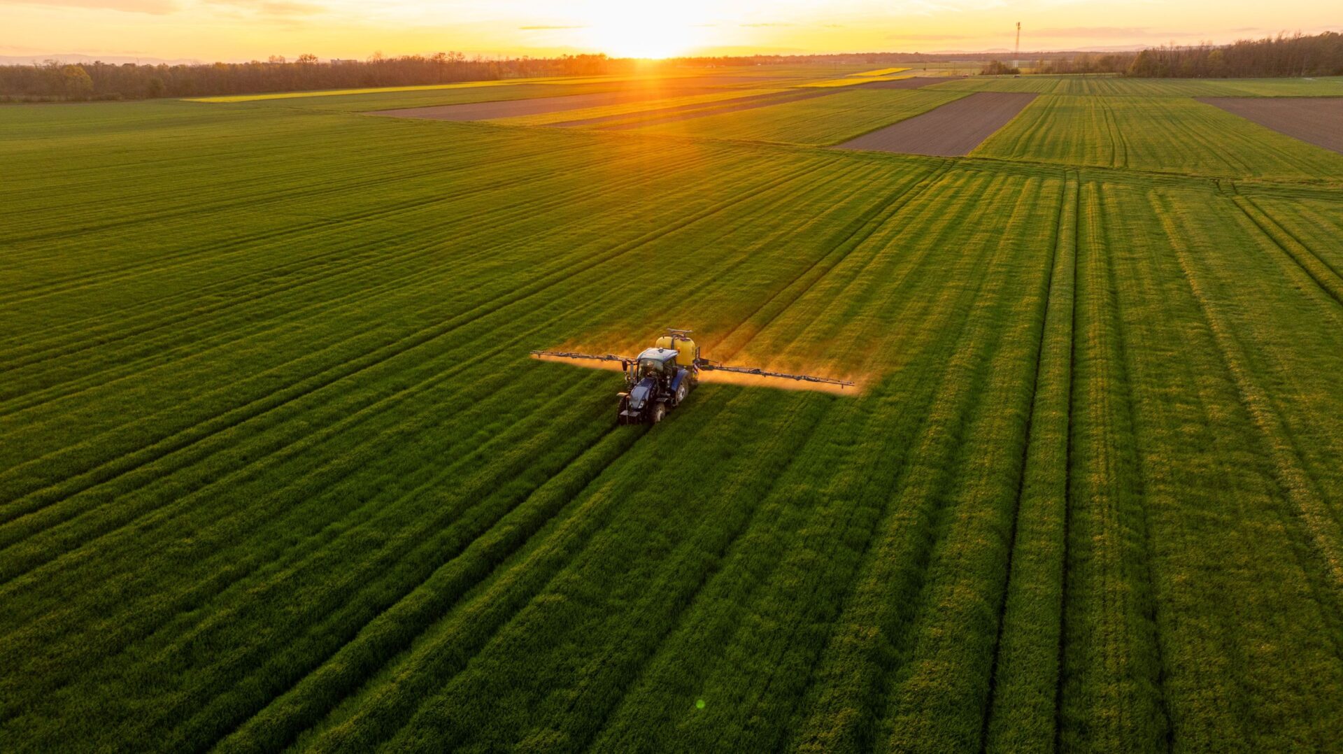 Aerial view of a tractor with a boom sprayer spraying crops across wide, green fields at sunset/sunrise.